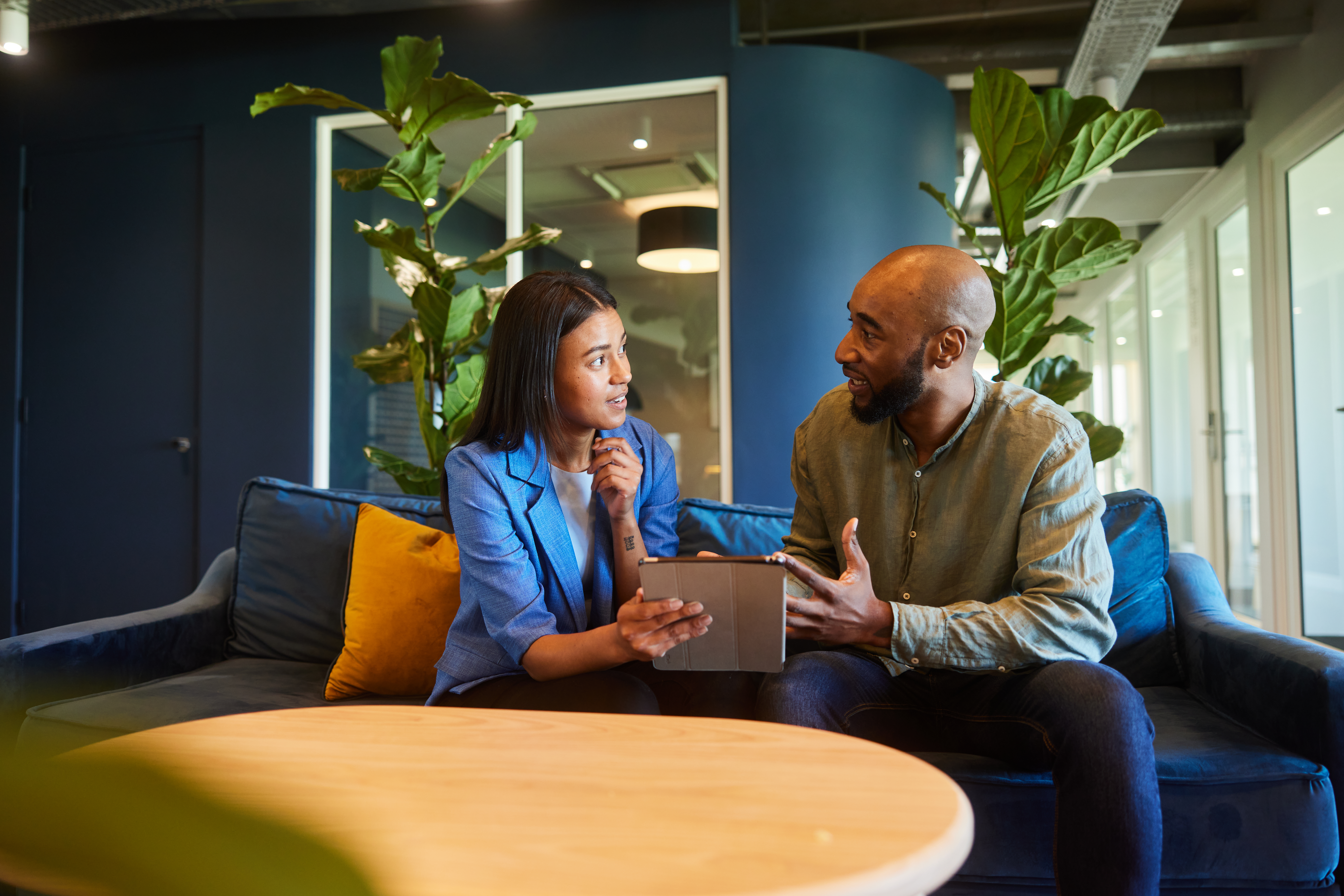Two real estate agents talking together over a digital tablet while sitting on a sofa in an office lounge